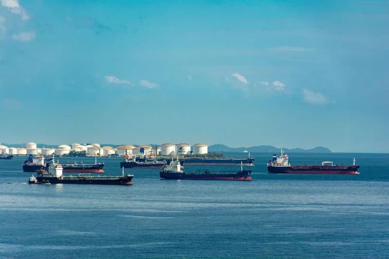 Cargo ships anchored beside oil storage tanks near a shipping lane, illustrating crude oil and the Strait of Hormuz.